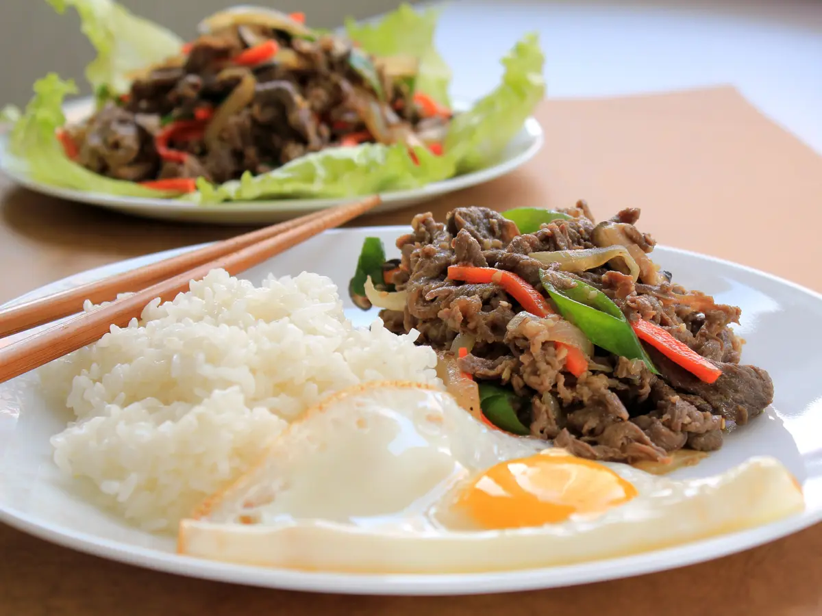 Beef bulgogi stir-fried with onion and colorful vegetables, served with white rice and a fried egg, with a second plate of bulgogi on lettuce leaves behind it
