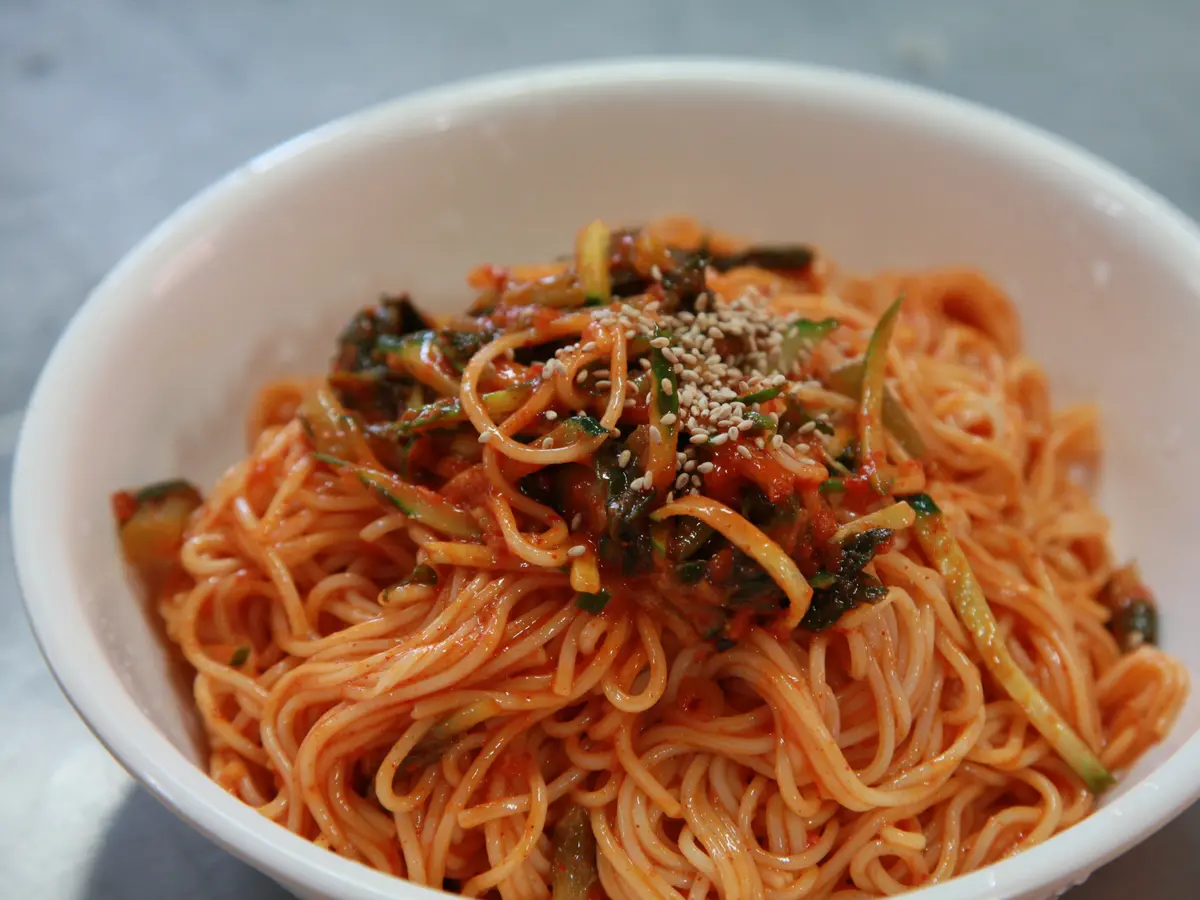 Bibim guksu in a white bowl: thin noodles coated in red chili sauce, topped with cucumber strips, chopped dark greens and sesame seeds