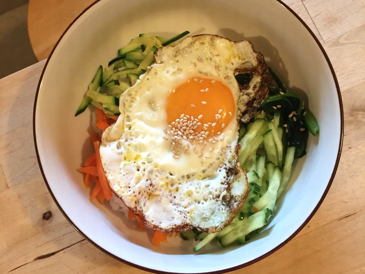 Bibimbap in a white bowl topped with a sunny-side-up fried egg and sesame seeds, surrounded by julienned cucumber, carrot and greens