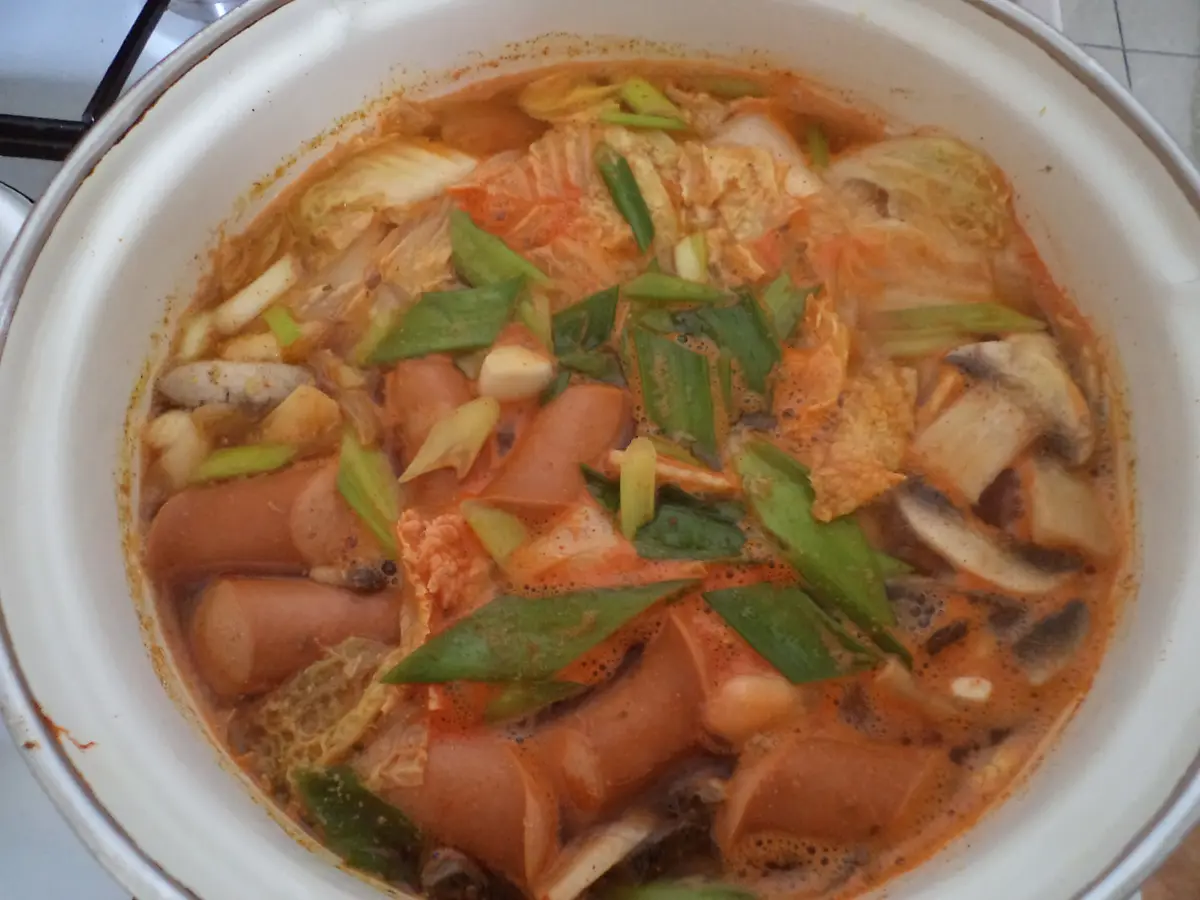 Budae jjigae army stew with sausage slices, kimchi, mushrooms and green onions in a white pot