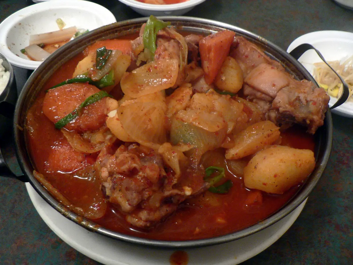 Dakdoritang chicken pieces with potato chunks, carrot slices, onion and green onion in red broth in a shallow metal pan, with small bowls of side dishes at the edges