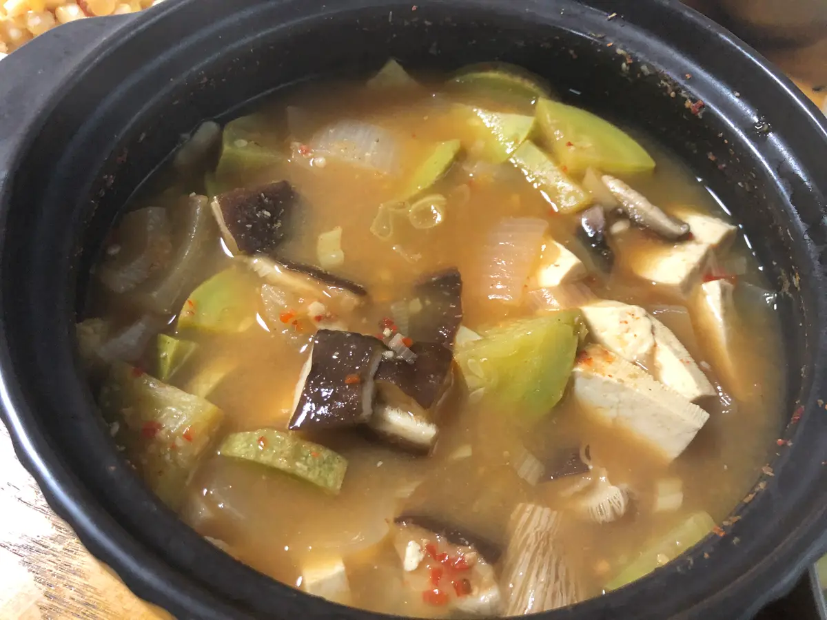 Doenjang jjigae in a black clay pot with cubed tofu, zucchini, sliced shiitake mushrooms and onion in a brown soybean paste broth flecked with red chili