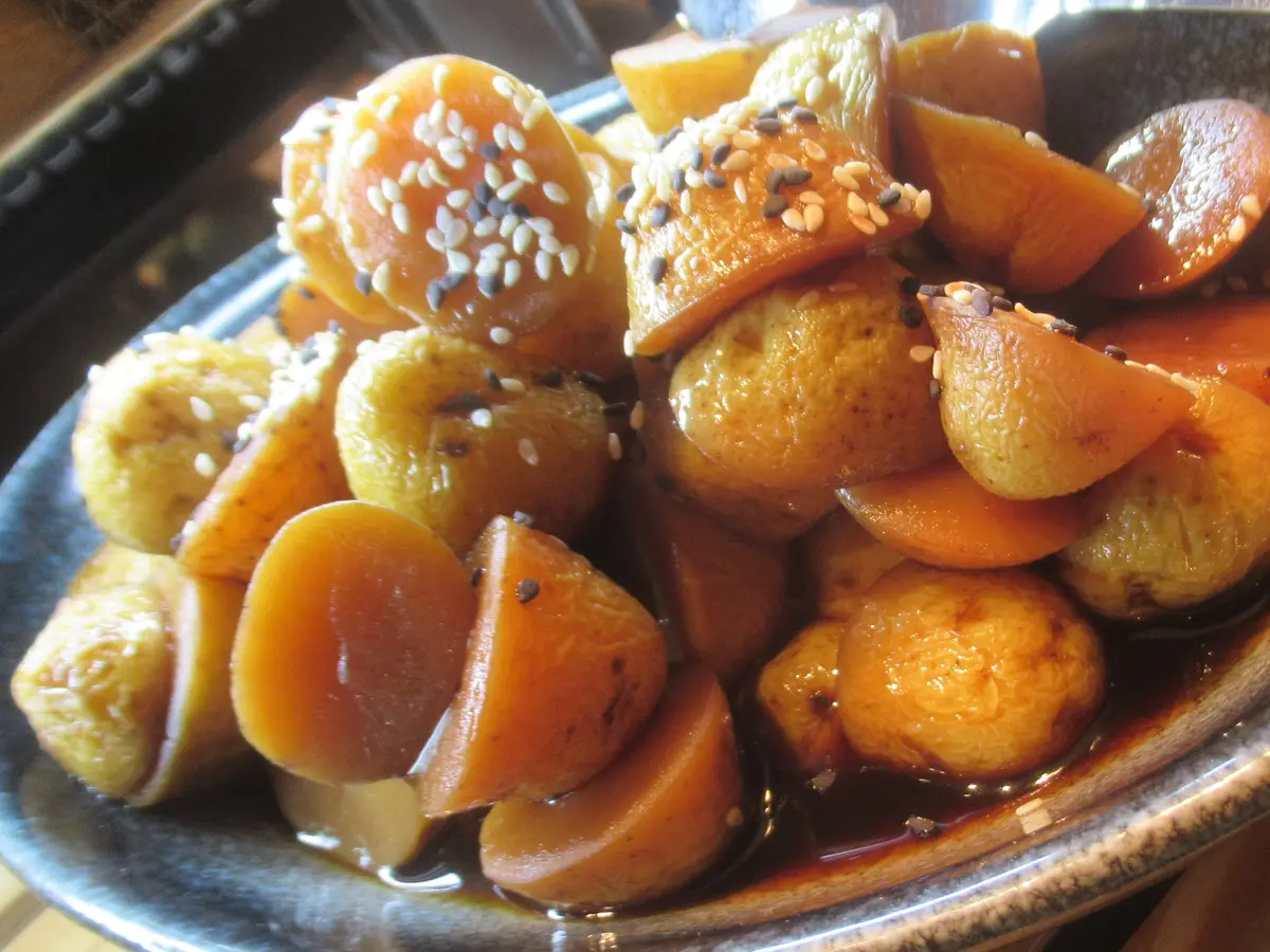 Close-up of gamja jorim: small whole potatoes and orange carrot slices in a glossy brown glaze with sesame seeds, in a gray-blue ceramic dish