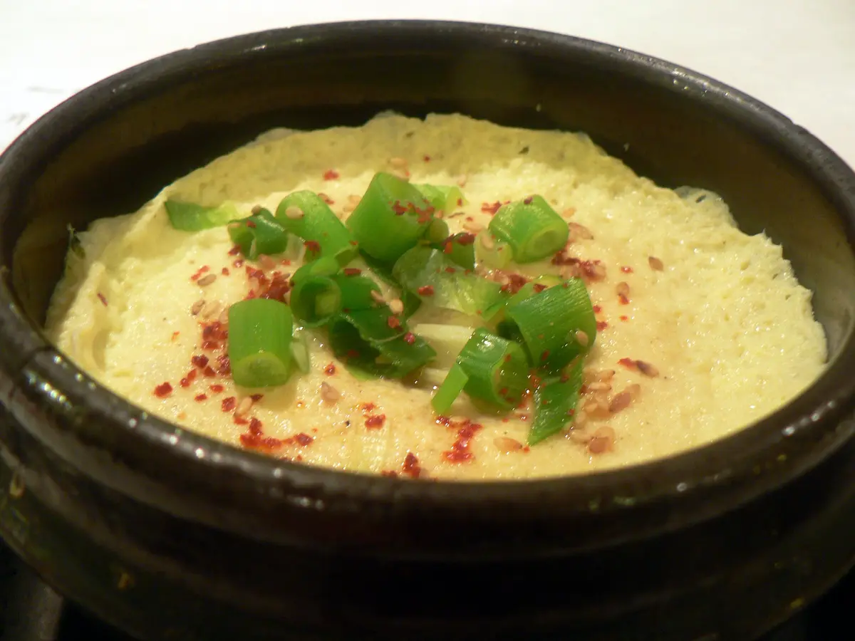 Korean steamed egg (gyeran jjim) puffed up in a black pot, topped with sliced green onions, sesame seeds and red pepper flakes
