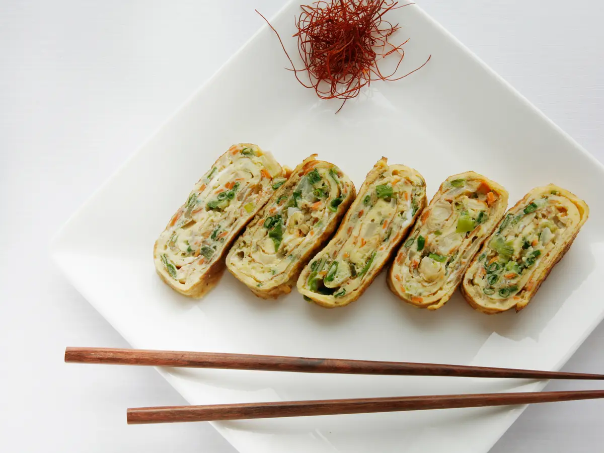 Sliced Korean rolled omelette with scallion and carrot spirals on a white plate, with wooden chopsticks and a garnish of red chili threads