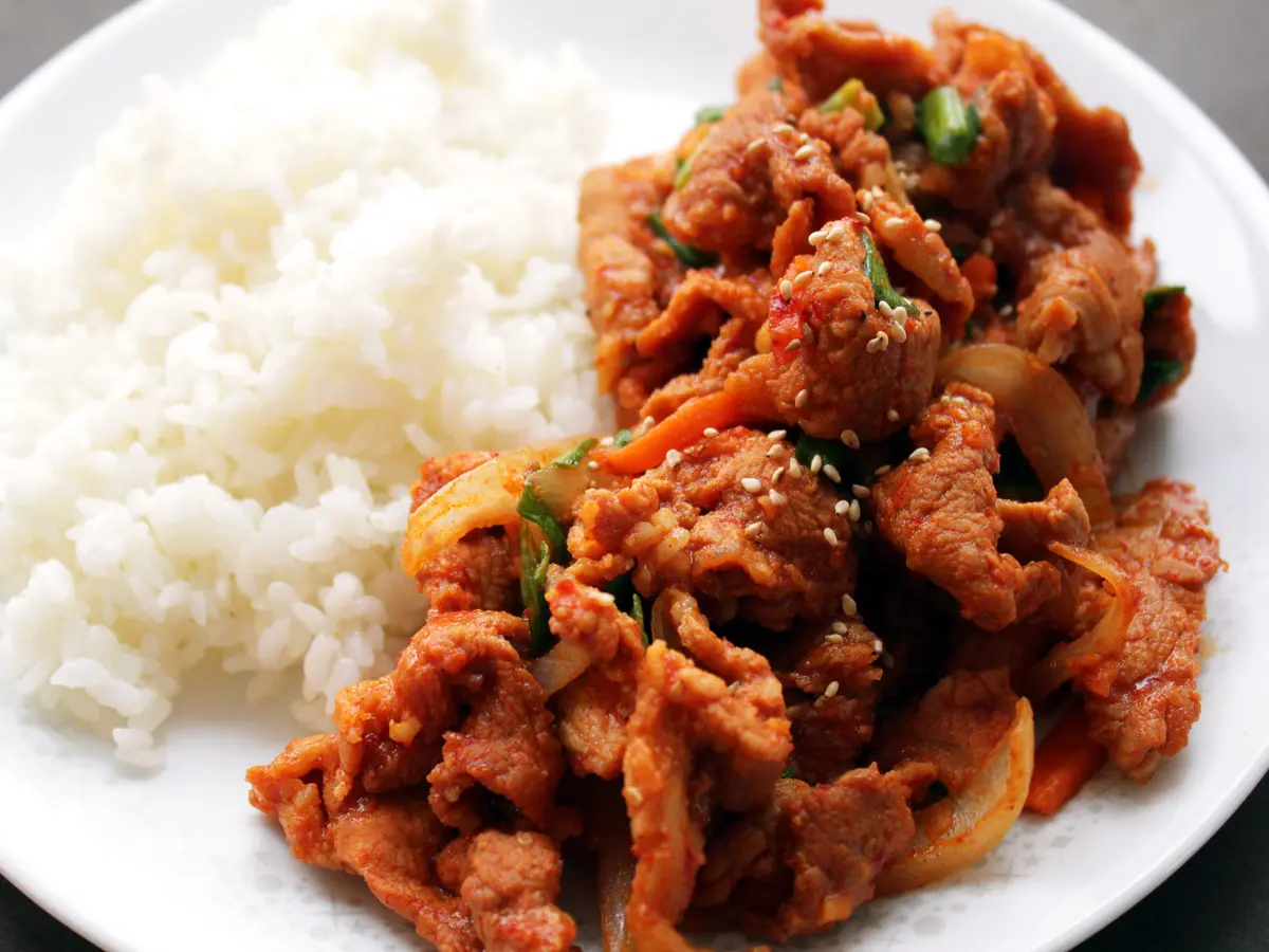 Spicy Korean stir-fried pork slices in a glossy red chili sauce with onion, carrot and scallions, topped with sesame seeds, beside white rice on a white plate