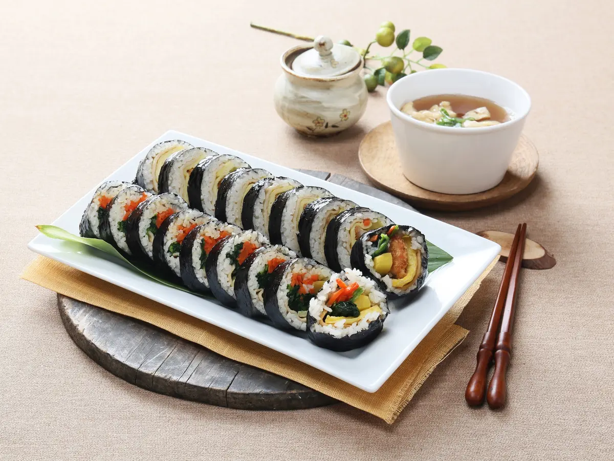 Sliced kimbap rolls on a white rectangular plate with a bowl of soup and wooden chopsticks