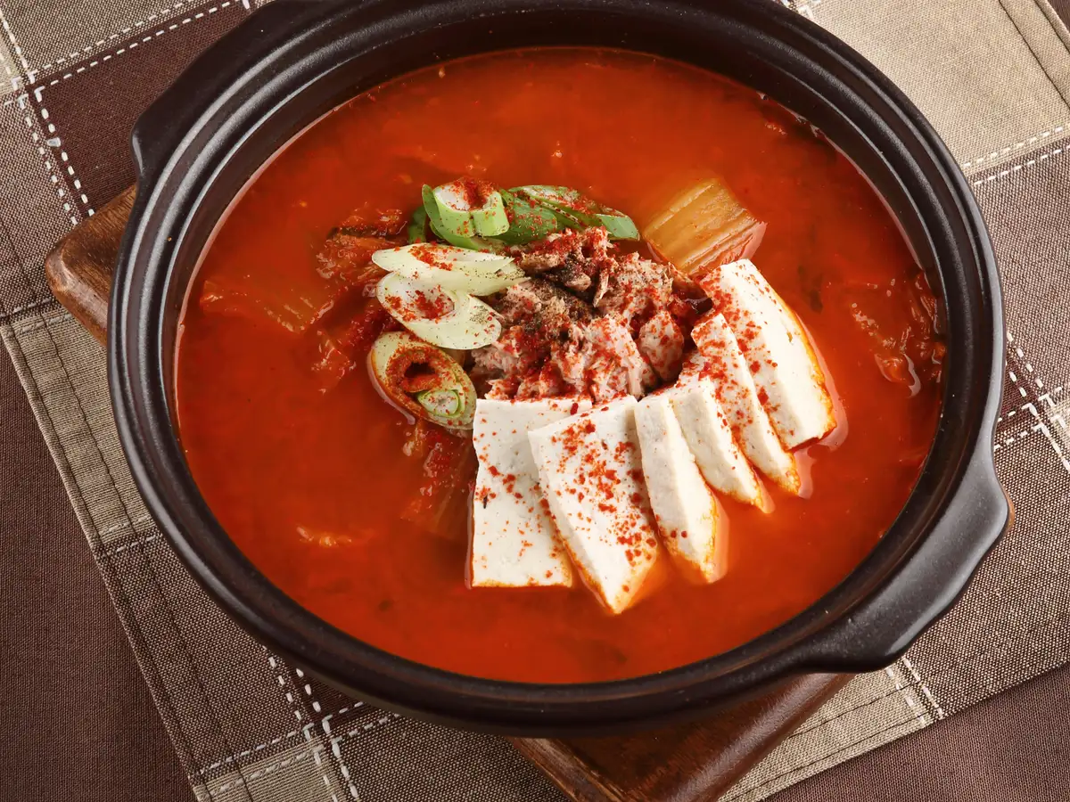 Kimchi jjigae bubbling in a black earthenware pot, topped with sliced tofu, pork, and green onion