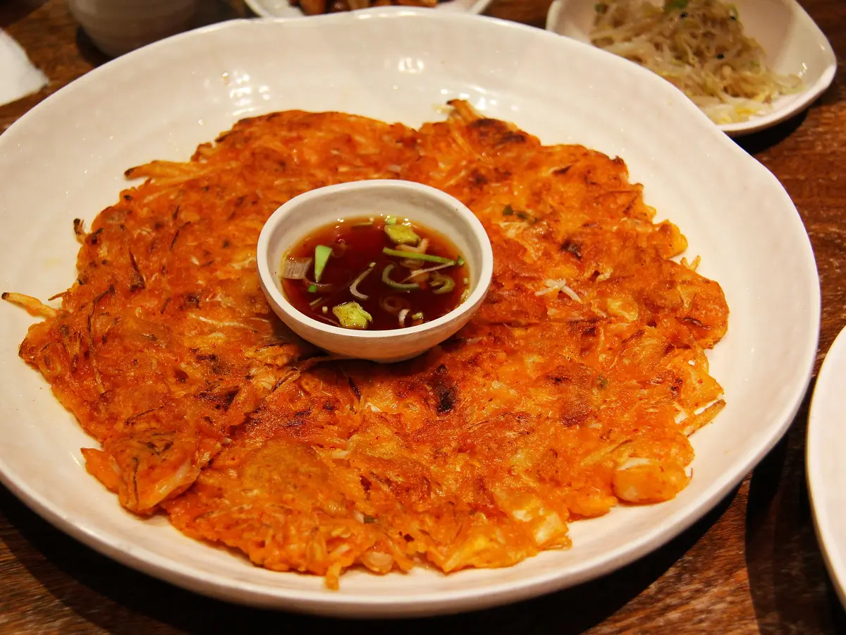 Crispy orange kimchi pancake on a white plate with a small dish of soy dipping sauce in the center