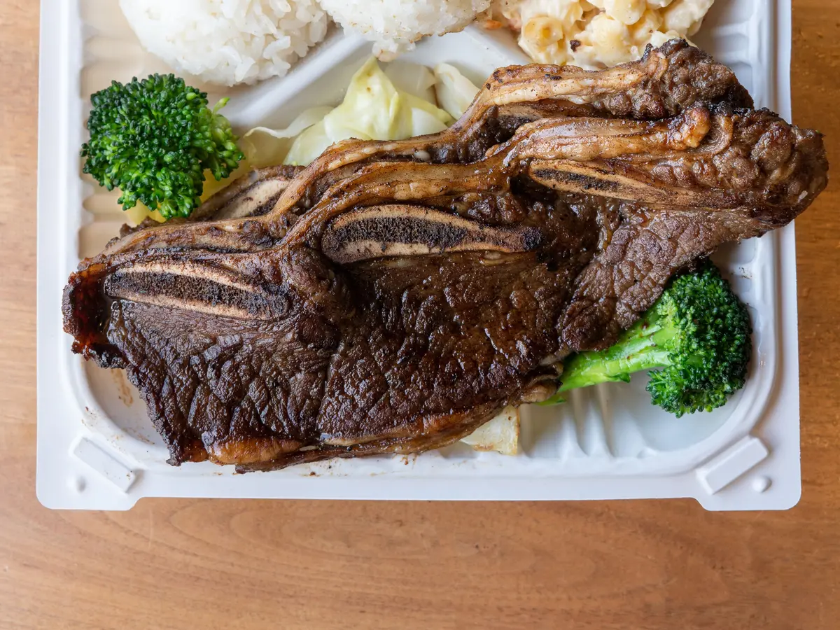Grilled LA galbi, flanken-cut beef short ribs with cross-cut bone slices, in a white takeout container with broccoli, cabbage, rice and macaroni salad