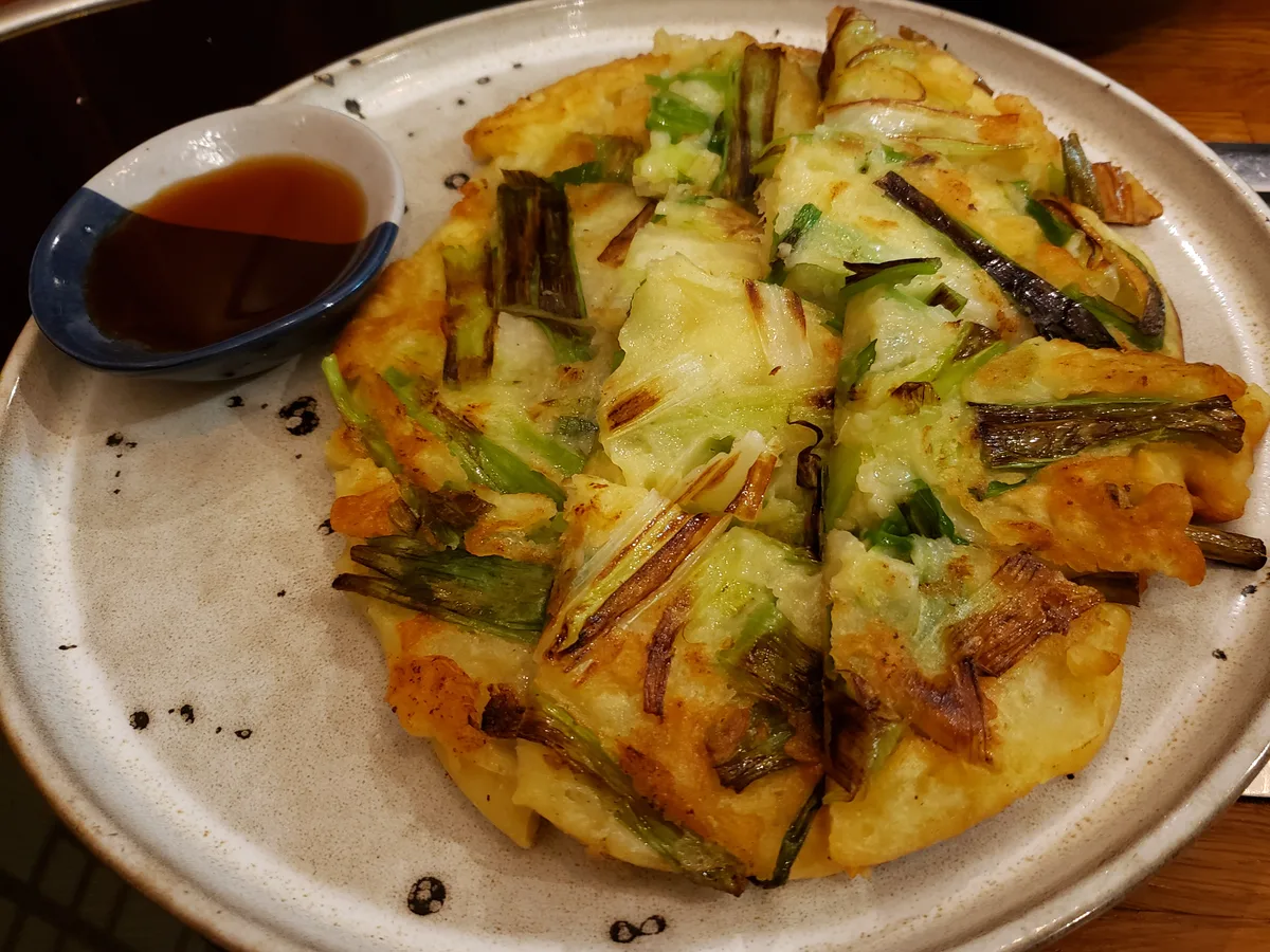 Golden scallion pajeon cut into wedges on a speckled ceramic plate with a small dish of soy dipping sauce