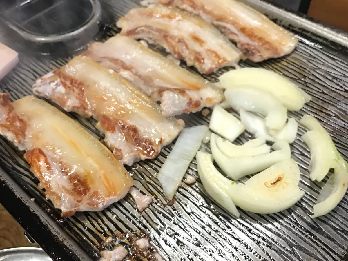 Grilled strips of pork belly showing layers of fat and meat, next to sliced onion, on a black ridged grill plate