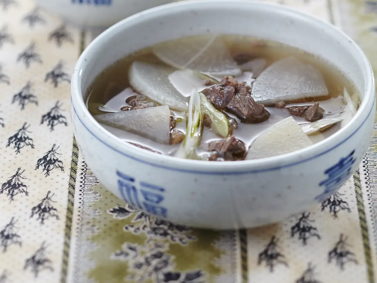 Soegogi muguk (Korean beef and radish soup) in a white bowl with blue characters on a patterned cloth, with sliced white radish, pieces of meat and green onion in a light brown broth