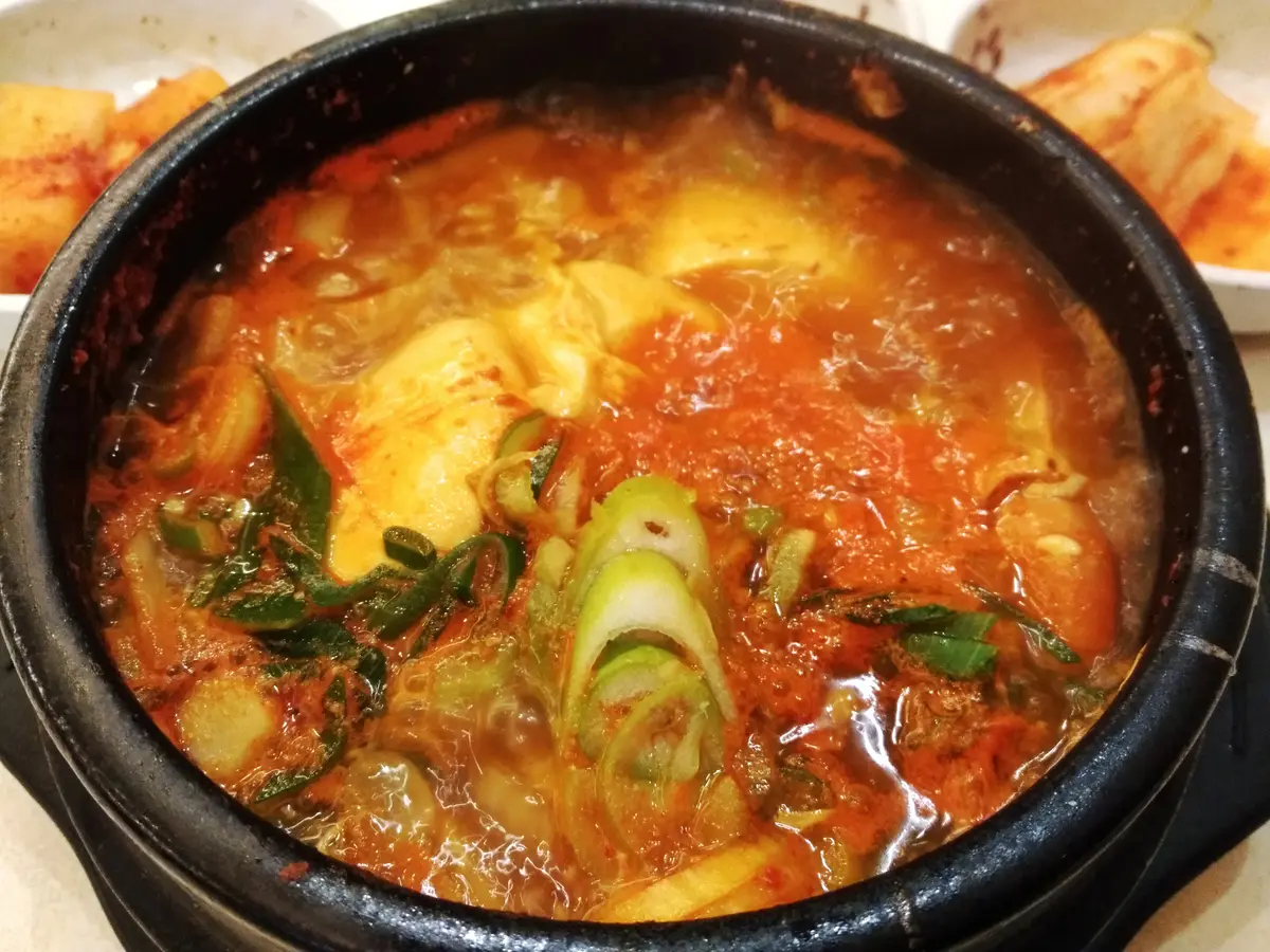 Bubbling red sundubu jjigae with soft tofu curds and sliced scallions in a black earthenware pot, with kimchi side dishes behind