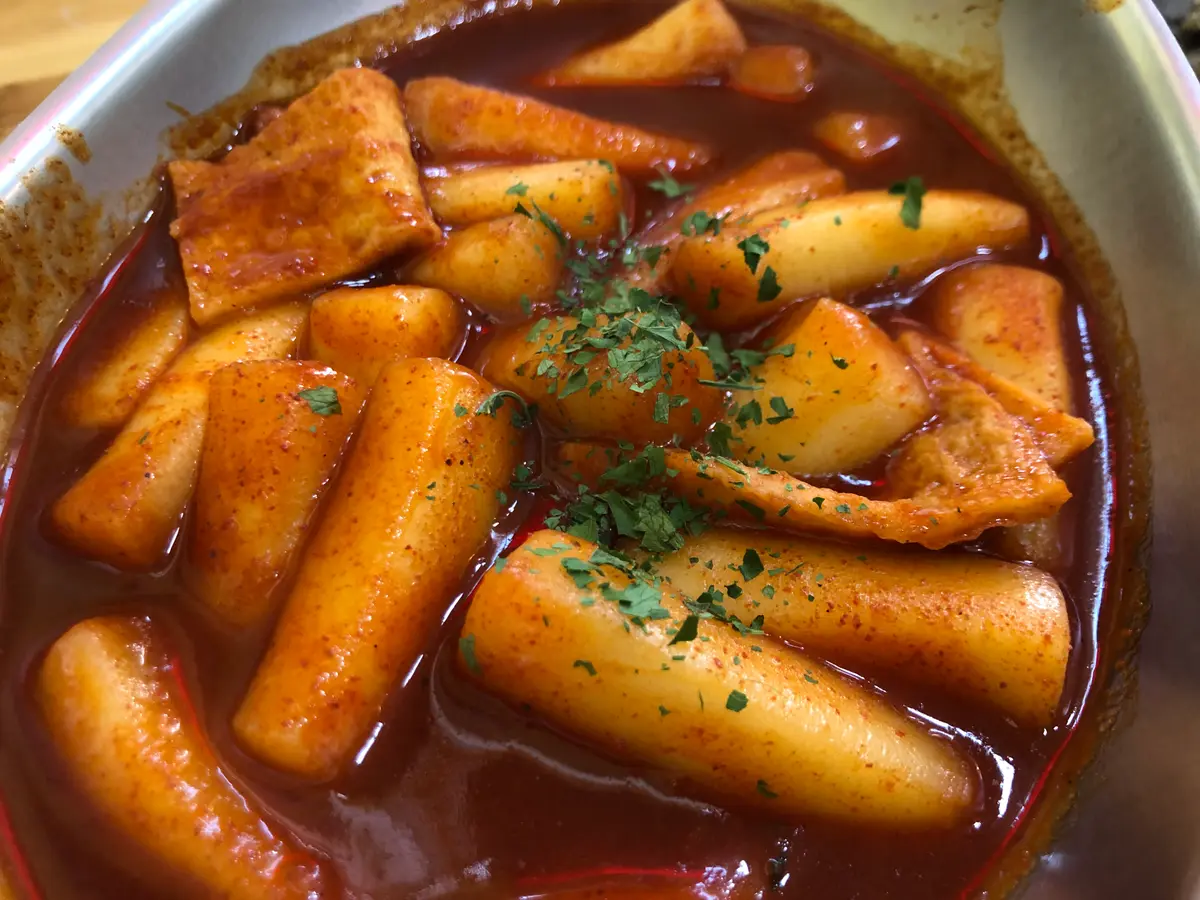 Close-up of tteokbokki: cylindrical rice cakes and fish cake pieces in a glossy red chili sauce, sprinkled with dried parsley flakes